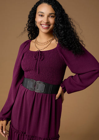 A woman modeling a long-sleeve smocked dress in deep plum purple with a wide black belt, set against a solid warm brown studio background.