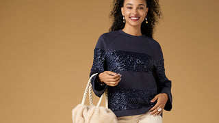 A woman modeling a navy blue crewneck sweater with horizontal sequined panels across the chest and sleeves, paired with beige pants and holding a cream-colored tote bag against a solid tan background.