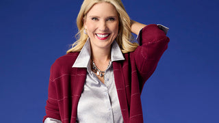 A smiling blonde woman posing against a solid blue background, wearing a maroon windowpane plaid cardigan over a shiny silver satin blouse.